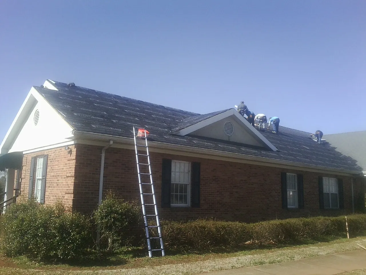 Crew tearing shingles off a brick ranch roof with a ladder set — Emergency Roof Repair work by our crew in Hanna