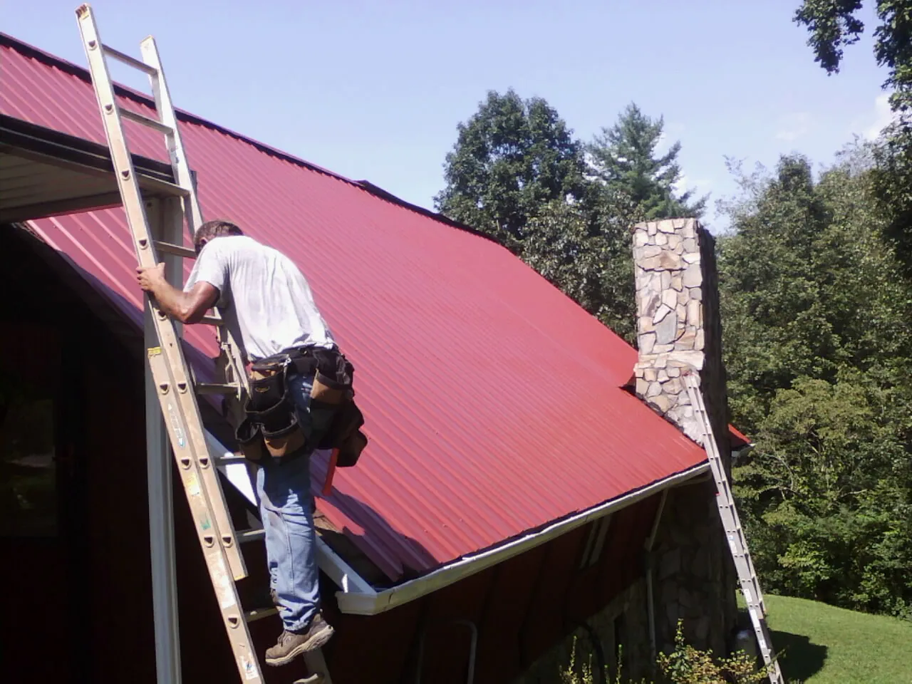 Roofer on a ladder beside a red metal roof and stone chimney — Soffit & Fascia Repair by Roofing Brothers in Hanna